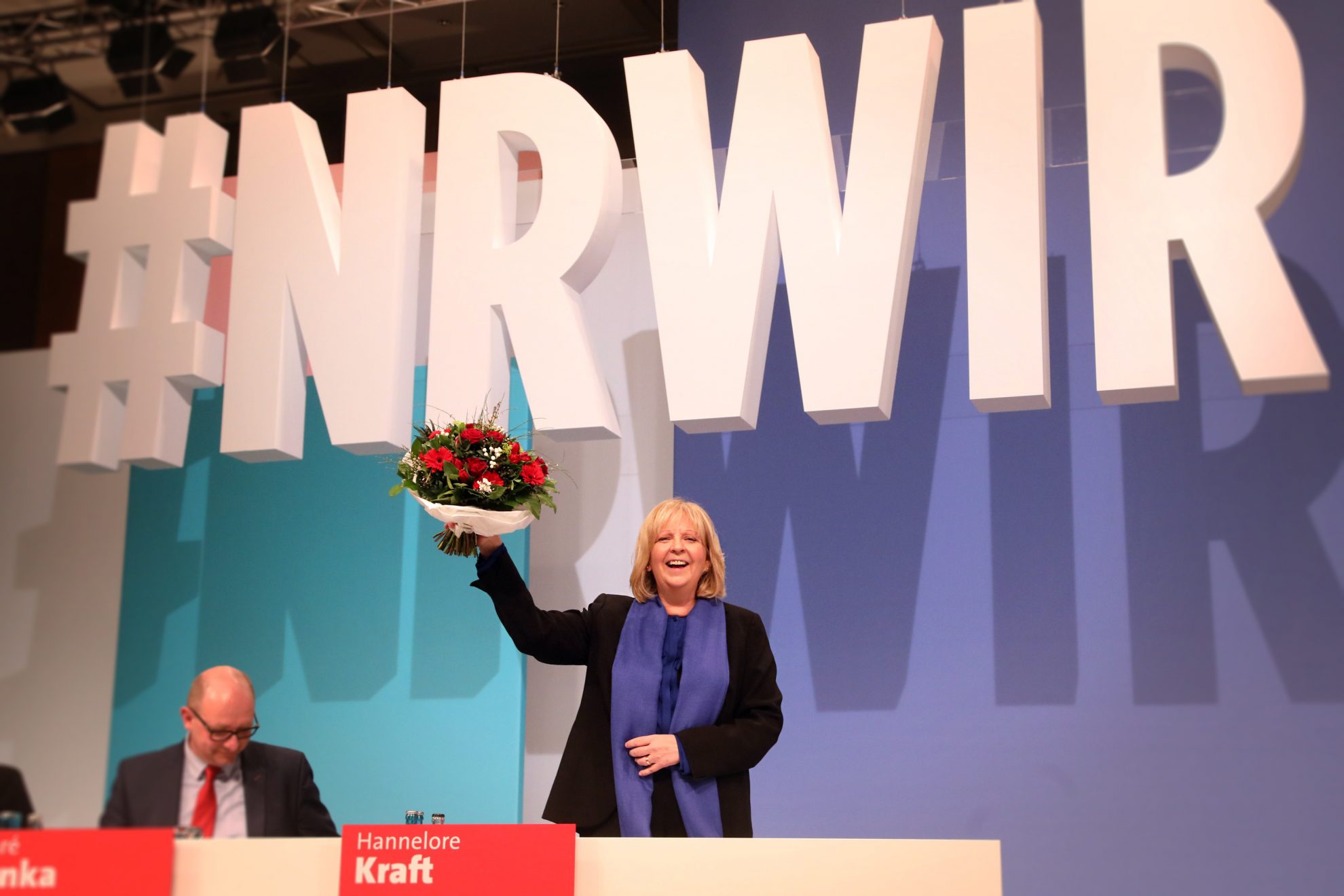 Hannelore Kraft steht lachend mit einem Blumenstrauß in der Hand auf dem Podium bei #NRWIR