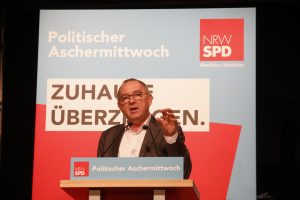 Norbert Walter-Borjans steht am Rednerpult mit erhobener Hand. Im Hintergrund ein Aufsteller in Blau und Rot mit der Aufschrift Politischer Aschermittwoch und "Zuhause überzeugen". Dem Slogan der bevorstehenden Kommunalwahl in NRW
