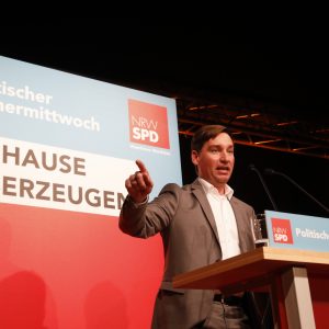 Sebastian Hartmann steht am Rednerpult mit erhobener Hand. Im Hintergrund ein Aufsteller in Blau und Rot mit der Aufschrift Politischer Aschermittwoch und "Zuhause überzeugen". Dem Slogan der bevorstehenden Kommunalwahl in NRW